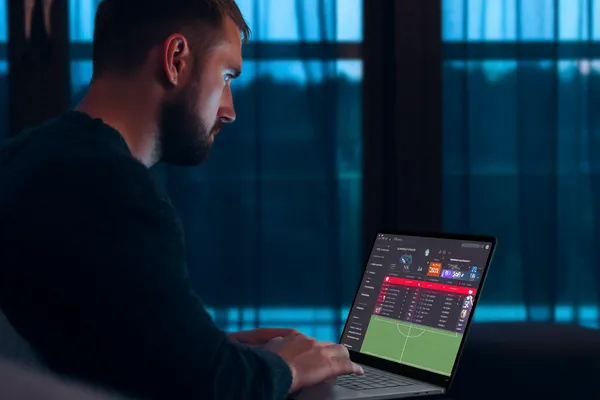 একজন পুরুষ তার ল্যাপটপে মনোযোগ দিয়ে কাজ করছেন, ব্যাকগ্রাউন্ডে লাইভ স্ট্যাটস — যা GRAPWIN মোবাইল অ্যাপের স্পোর্টস ও গেমিং ফিচারগুলোকে তুলে ধরে।